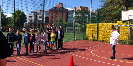 Powiększ grafikę: Uczniowie stoją w rzędzie na boisku do koszykówki. W tle widać budynki. Po prawej stronie znajduje się nauczycielka.