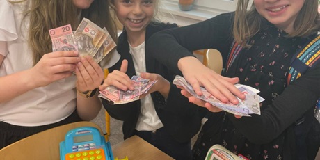 Powiększ grafikę: Trzy uśmiechnięte dziewczynki z banknotami w ręce. Na ławce leżą pieniądze i stoją dwie kasy