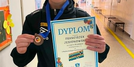 Powiększ grafikę: Chłopiec stoi na korytarzu z dyplomem i medalem w ręku za 3 miejsce w Międzyszkolnym Turnieju Kostki Rubika