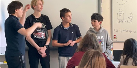 Powiększ grafikę: 4 uczniów stojących na tle białej tablicy multimedialnej.   Jeden uczeń w granatowej koszulce wymachuje rękoma, próbując w ten sposób tłumaczyć.