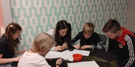 Powiększ grafikę: 3 chłopców i 2 dziewczynki siedzące przy okrągłym stole, piszące alfabetem Braille’a. Z tylu zielona ściana