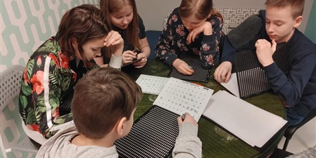Powiększ grafikę: 2 chłopców i 3 dziewczynki siedzące przy okrągłym stole, piszące alfabetem Braille’a. Z tylu zielona ściana