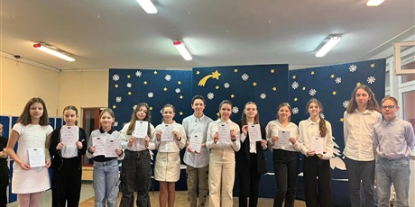 Powiększ grafikę: 12 dzieci stoi na scenie i pokazuje swoje dyplomy. Za nimi niebieska tablica z gwiazdkami i śnieżynkami. 