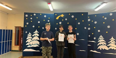 Powiększ grafikę: 3 dzieci stoi na scenie i pokazuje swoje dyplomy. Za nimi niebieska tablica z gwiazdkami i śnieżynkami. 