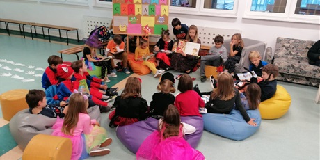 Powiększ grafikę: Uczniowie siedzą na pufach i kanapie i słuchają, jak nauczycielka czyta im książkę. W te widać napis: Strefa Czytania.
