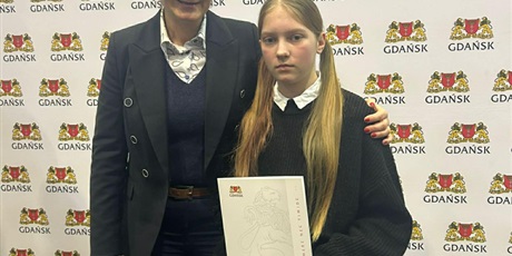 Powiększ grafikę: Dziewczynka stoi u boku pani prezydent Gdańska z dyplomem w ręce  w tle tablica z herbem Gdańska