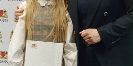 Powiększ grafikę: Dziewczynka stoi u boku pani prezydent Gdańska z dyplomem w ręce  w tle tablica z herbem Gdańska