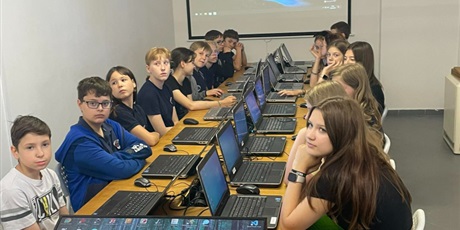 Powiększ grafikę: Grupa uczniów siedząca przy komputerze. W tle tablica multimedialna
