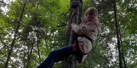Powiększ grafikę: wśród drzew na środku zdjęcia dziewczynka wspinającą się po linie na czubek drzewa. Na zielono napisane słowo Karolina