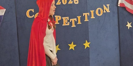 Powiększ grafikę: Dziewczynka ubrana w czerwoną pelerynę z kapturem recytuje wiersz o wilku, babci i Czerwonym Kapturku