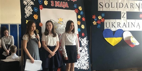 Powiększ grafikę: 3 uczennice recytują wiersze. W tle widać napis: Konstytucja 3 maja i solidarni z Ukrainą. Za sceną uczniowie, którzy czekają na swój występ.